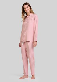 Mujer sonriente de pie descalza, vistiendo una blusa de pijama de manga larga a rayas en rosa claro y pantalones a juego, contra un fondo sencillo.
