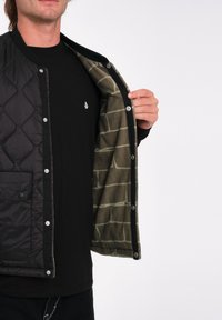 Chaqueta negra acolchada con botones de presión, con un forro verde estampado. Llevada sobre una camisa negra con un pequeño logotipo en el pecho.