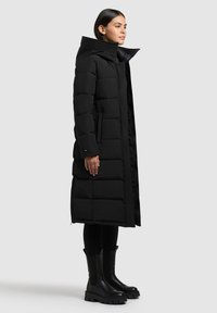 Schwarze Daunenjacke mit hohem Kragen, mit einem gesteppten Design, Fronttaschen und einer eleganten, langen Silhouette. Kombiniert mit schwarzen Knöchelstiefeln.