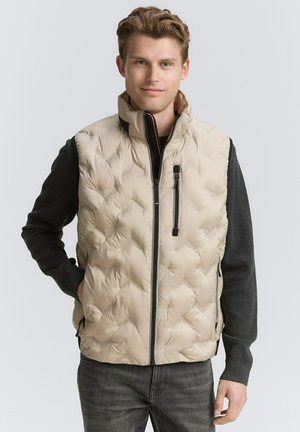 Chaleco puffer beige con un diseño acolchado, cuello alto y cremallera frontal. Cuenta con bolsillos laterales y un suéter oscuro en contraste debajo.