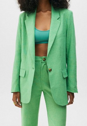 Personne portant un costume vert clair avec un blazer et un pantalon taille haute, associé à un top court côtelé de couleur sarcelle.