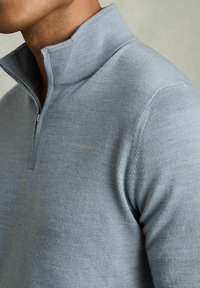 Pulover gri cu jumătate de fermoar, realizat dintr-un material tricotat moale, având un guler înalt, accente ribbed și o formă ajustată. Textură netedă cu un luciu subtil.