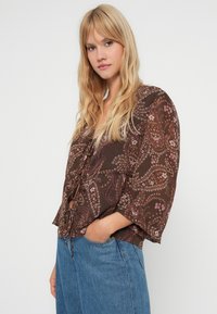 Rjava cvetlična bluzica z razširjenimi rokavi, ki ima vrvico na pasu in zapletene paisley vzorce v roza in belem.