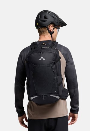Zaino nero multifunzionale con compartimenti, spallacci imbottiti, tasche in rete e dettagli riflettenti. Indossato da una persona con un casco sportivo.