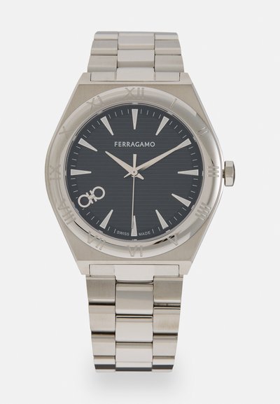 Reloj de acero inoxidable plateado con un dial texturizado gris oscuro, marcadores de hora plateados y una pulsera de eslabones. Presenta números romanos y un logotipo.