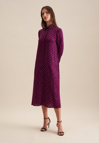 Vestito a maniche lunghe di un vivace magenta con un motivo geometrico ripetuto. Tessuto liscio, chiusura con bottoni davanti e lunghezza a metà polpaccio. Tacchi neri.