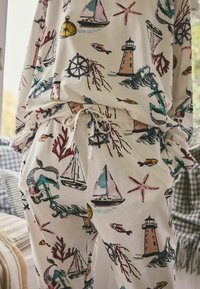 Bavlněné domácí oblečení s námořnickým potiskem zobrazujícím plachetnice, majáky a mořské prvky v červené, modré a růžové barvě na krémovém pozadí.
