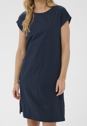 Mujer con un vestido sencillo azul marino de manga corta con puños remangados y pulseras doradas en ambas muñecas.