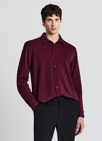 Bordeauxrotes Langarm-Hemd mit Knopfleiste, strukturiertem Stoff mit dezentem Schimmer, klassischem Kragen und spitzen Manschetten, kombiniert mit schwarzen Hosen.