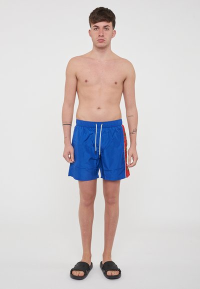Shorts de bain bleus avec un panneau latéral rouge, taille élastique et cordon de serrage. Portés avec des sandales noires, le fond est uni blanc.