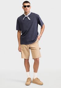 Mann mit schwarzen Sonnenbrillen, dunkelgraßem übergroßem T-Shirt, beigen Shorts, beigen Turnschuhen, weißen Socken und einem weißen Halstuch, der mit einer Hand in der Tasche steht.