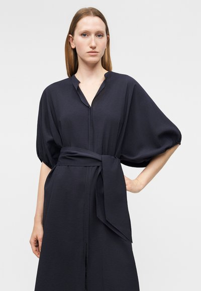 Γυναίκα με ίσια ανοιχτόκασταμα καστανά μαλλιά που φοράει μαύρο φόρεμα με φαρδιά μανίκια και ζώνη, στέκεται μπροστά σε απλό ανοιχτό φόντο.