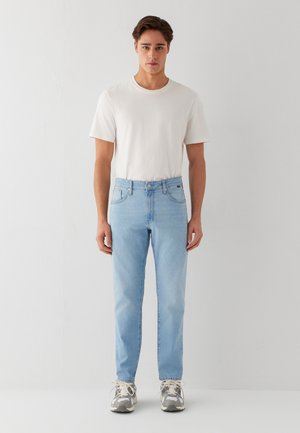 Junger Mann trägt ein schlichtes weißes T-Shirt, hellblaue Jeans und graue Sneaker und steht vor einem weißen Hintergrund.