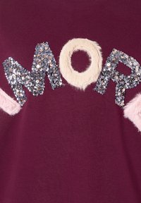 Nahaufnahme von burgunderfarbenem Stoff, auf dem das Wort 'MORE' in silbernen Pailletten und mit crem farbenem, flauschigem Material auf den Buchstaben geschrieben ist.