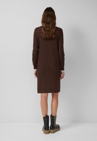 Robe marron à manches longues avec une coupe droite, arrivant au genou, et un détail de poignet blanc. Associée à des bottines noires à chevilles avec des côtés texturés.