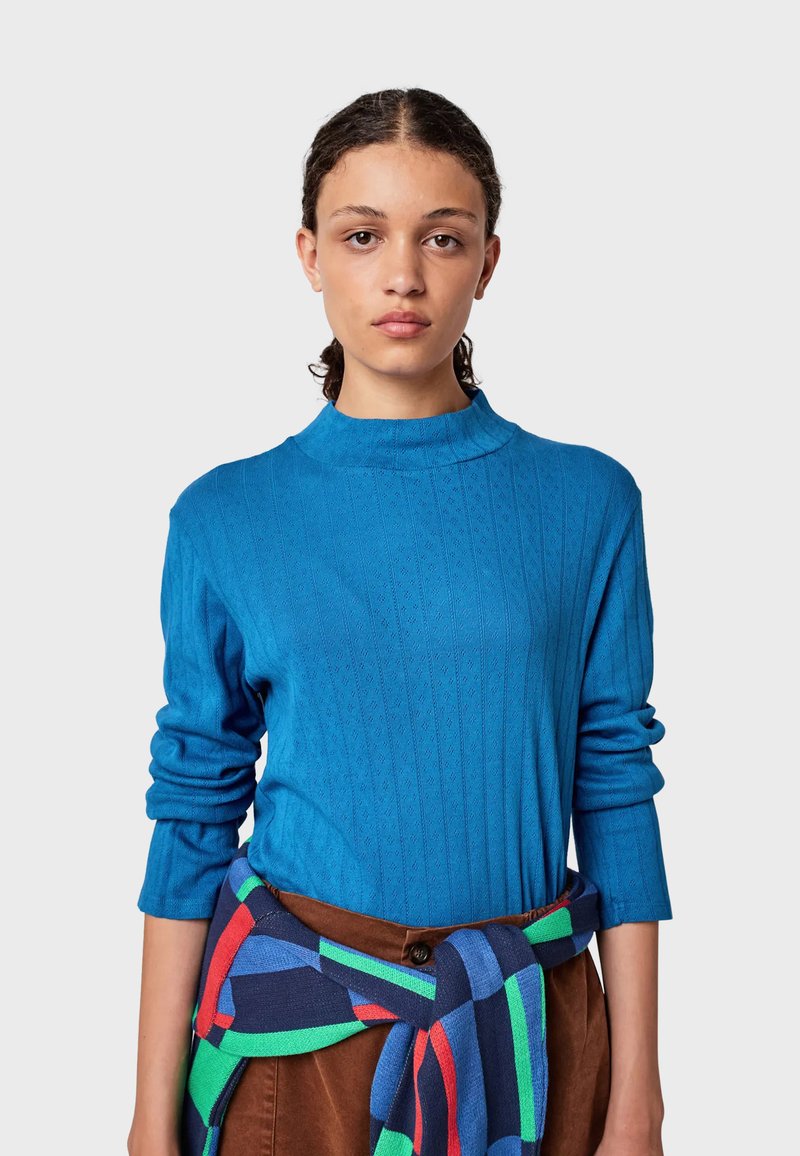 Maglietta blu a maniche lunghe con colletto alto e design a costine. Modella che indossa una gonna marrone con una sciarpa a fantasia multicolore legata in vita.