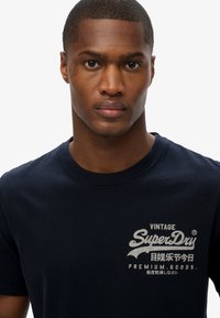 Marineblauw katoenen T-shirt met een "SuperDry"-logo in het wit op de borst, een ronde hals en korte mouwen.