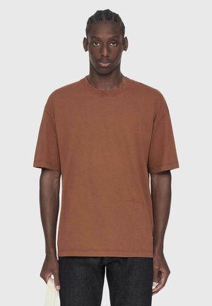 Hombre con el pelo trenzado que lleva una camiseta marrón de gran tamaño y pantalones negros, de pie y sosteniendo un paño claro doblado en su mano izquierda.