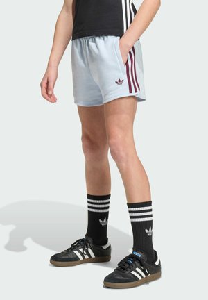 Persona vistiendo pantalones cortos Adidas azul claro con rayas marrones, calcetines Adidas negros y zapatillas Adidas negras con rayas blancas.