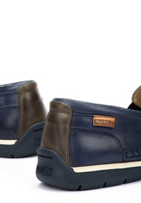 Zapatos de cuero azul oscuro y oliva con suela de goma, que presentan un diseño cosido y una etiqueta de marca en la parte trasera. Forma redondeada, estilo bajo.
