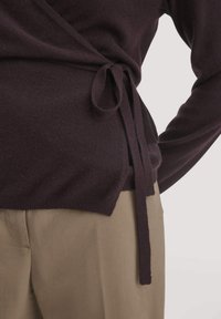 Dunkel-lila Wickeljacke mit langen Ärmeln, Taillenbund und weichem Material, kombiniert mit hellbeigen Hosen.