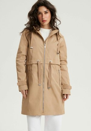 Kobieta z falującymi ciemnymi włosami, ubrana w beżowy długi płaszcz z kapturem, suwakiem z przodu i sznurkiem w talii, na białych spodniach.