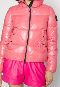 Chaqueta de plumas rosa con un acabado brillante, cierre de cremallera y dos bolsillos laterales con botones negros. Diseño de longitud corta y cuello alto.