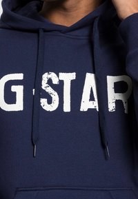 Osoba ve tmavě modré mikině s kapucí, na které je bílé logo "G-STAR" a viditelné stahovací šňůrky, zvedající kapuci u krku.