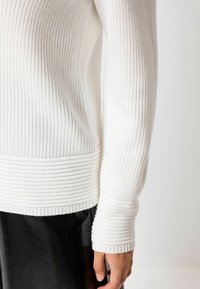 Pull en maille côtelée blanche avec un tissu texturé, des poignets ajustés et un motif rayé horizontal le long de la taille.