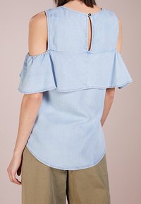 Kvinna klädd i en ljusblå blus med kalla axlar, frilldetaljer och nyckelhålsknappstängning på baksidan, tillsammans med olivgröna byxor.