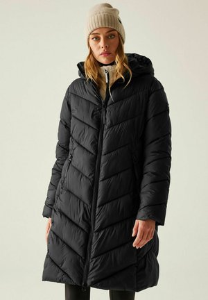 Kobieta w czarnej pikowanej, długiej puchowej kurtce z kapturem i beżowej dzianinowej czapce na jednolitym jasnym tle.