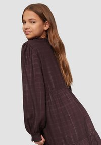 Bruine geblokte jurk met lange puffmouwen, een geplooide halslijn en een gelaagde rok. De stof heeft een subtiele textuur en glans.