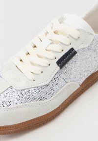 White sneaker with textured suede and shiny silver accents. Features cream laces, a black detail, and a brown rubber sole.