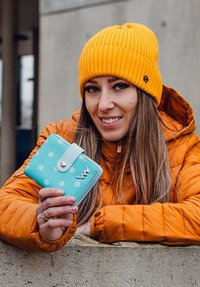 Light blue wallet with white polka dots and a snap closure, held by a person in an orange puffer jacket and yellow ribbed beanie.