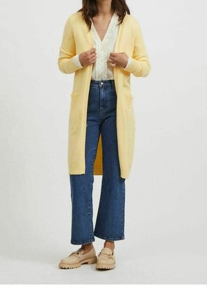 Kvinde iført en lang gul cardigan over en hvid bluse, blå jeans og beige loafers, stående mod en ensfarvet baggrund.