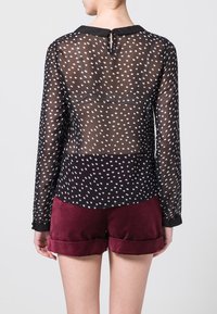 Blusa negra transparente con lunares blancos, mangas largas y cuello negro. Combinada con pantalones cortos burdeos con dobladillos enrollados.
