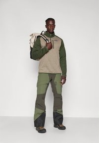 Maglione in pile con torso beige e maniche verdi, abbinato a pantaloni color oliva. Il modello porta uno zaino beige e indossa stivali neri.