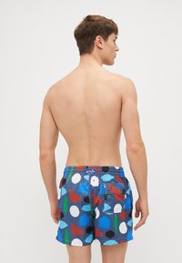 Μαγιό σε μπλε χρώμα με ένα μείγμα πολύχρωμων γεωμετρικών σχεδίων που περιλαμβάνουν κύκλους, τρίγωνα και φύλλα. Διαθέτει πίσω τσέπη και ελαστική ζώνη.