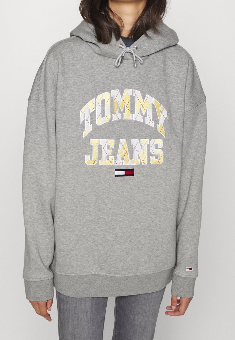 Personne portant un sweat à capuche gris avec le texte et le logo "TOMMY JEANS" sur le devant, associé à un jean gris, posant sur un fond uni.