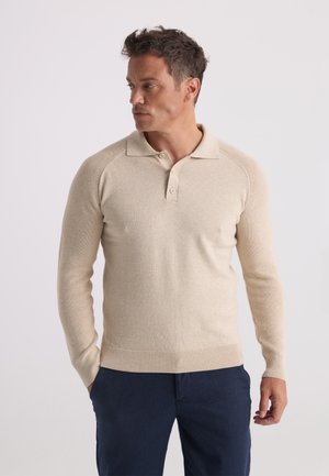 Homme portant un polo beige à manches longues et un pantalon bleu foncé, debout les mains dans les poches et regardant vers la gauche sur un fond uni.