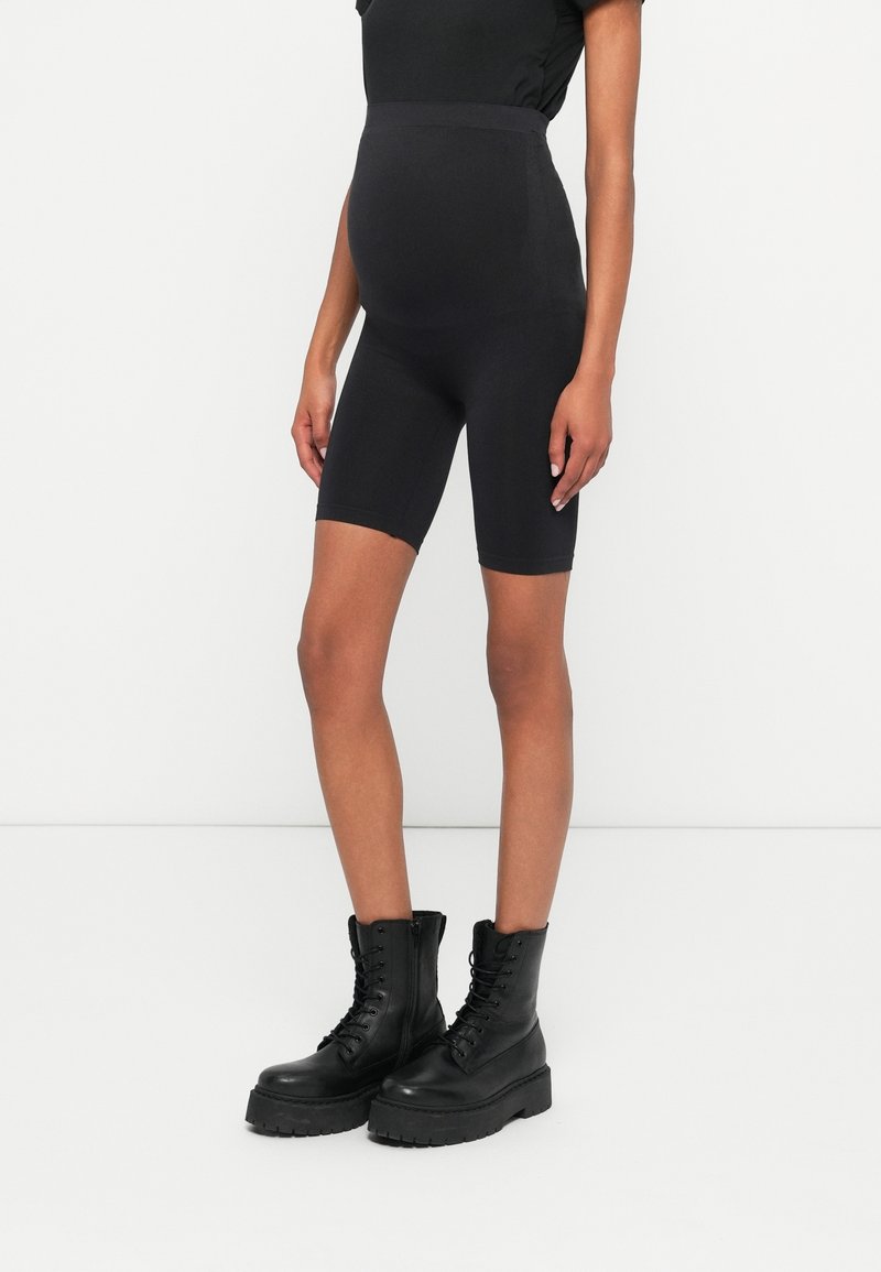 Femme enceinte portant un short biker noir taille haute et des bottes de combat noires à lacets, debout devant un fond blanc uni.