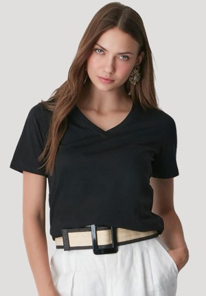 Junge Frau mit langen braunen Haaren, die ein schwarzes V-Ausschnitt-T-Shirt, große dekorative Ohrringe, weiße Hosen und einen beigen Gürtel mit schwarzer Schnalle trägt.