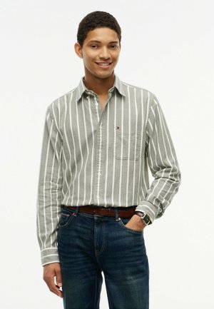 Ung mand smiler iført en grå og hvid vertikalt striped skjorte med knapper, mørke jeans og et brunt bælte, hånd i lommen.