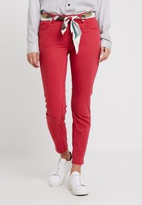 Jean rouge ajusté en denim avec une texture lisse, présentant une silhouette slim et des poches latérales, associé à des baskets blanches.
