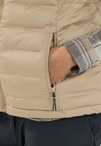 Beige vadderad väst med en dragkedjeficka, med ett rutigt skjortlager synligt vid ärmen och kontrasterande mörka byxor. Mjuk textur.