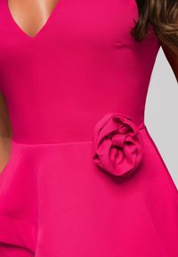 Lähikuva naisesta, jolla on päällään kirkkaan pinkki mekko, jossa on V-pääntie ja kangaskukka yksityiskohtana vyötäröllä vaaleaa taustaa vasten.