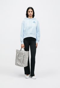 Ανοιχτό γαλάζιο cropped hoodie με μπροστινή τσέπη και λογότυπο, συνδυασμένο με μαύρο κολάν. Γκρι τσάντα tote με λογότυπο και χερούλι, φορεμένη με λευκά παπούτσια.