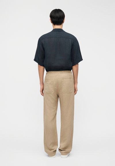 Άνδρας που στέκεται πλάτη, φοράει σκούρο κοντομάνικο πουκάμισο μέσα σε μπεζ παντελόνι με φαρδύ πόδι και πίσω τσέπες, και λευκά παπούτσια.