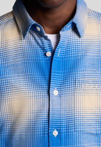 Button-up skjorte med et blå- og beige ternet mønster, lavet af bomuldsstof, standard krave og hvide knapper. Struktureret overflade med kontrastfarver.