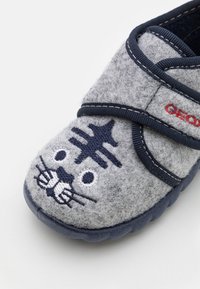 Szare kapcie z motywem kociej twarzy w niebieskim i białym haftem, z granatowym paskiem i gumową podeszwą zapewniającą komfort noszenia.